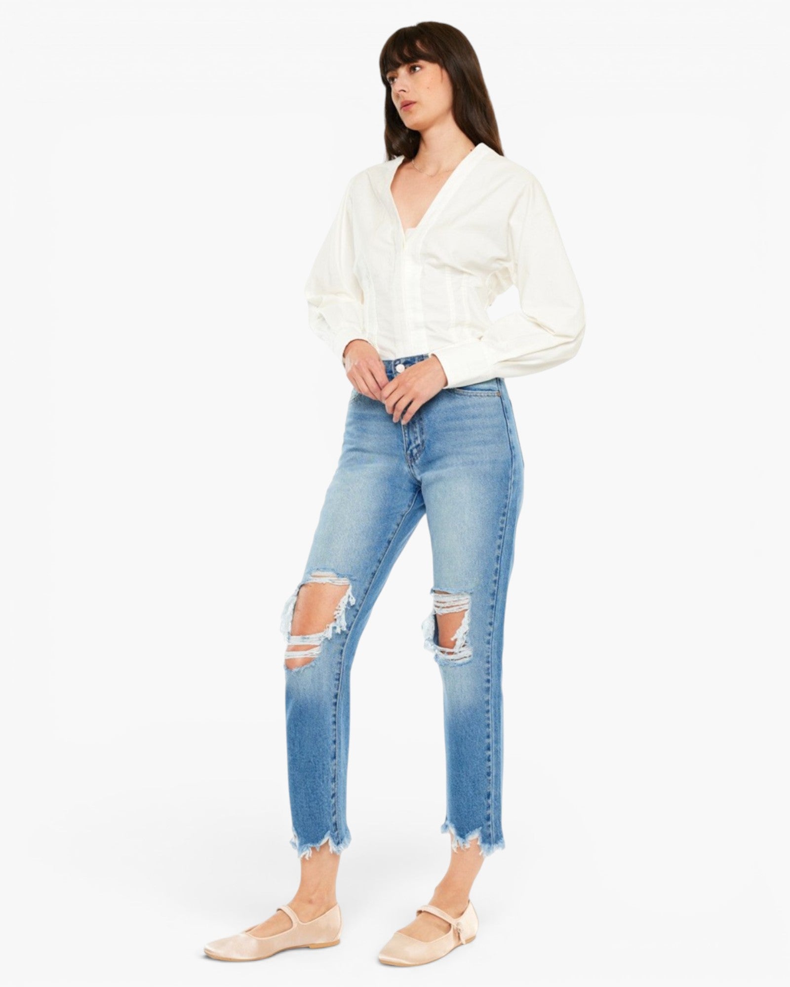 A model wearing light-wash blue distressed cropped jeans with frayed hems, paired with a white blouse and beige ballet flats, against a plain white studio background.