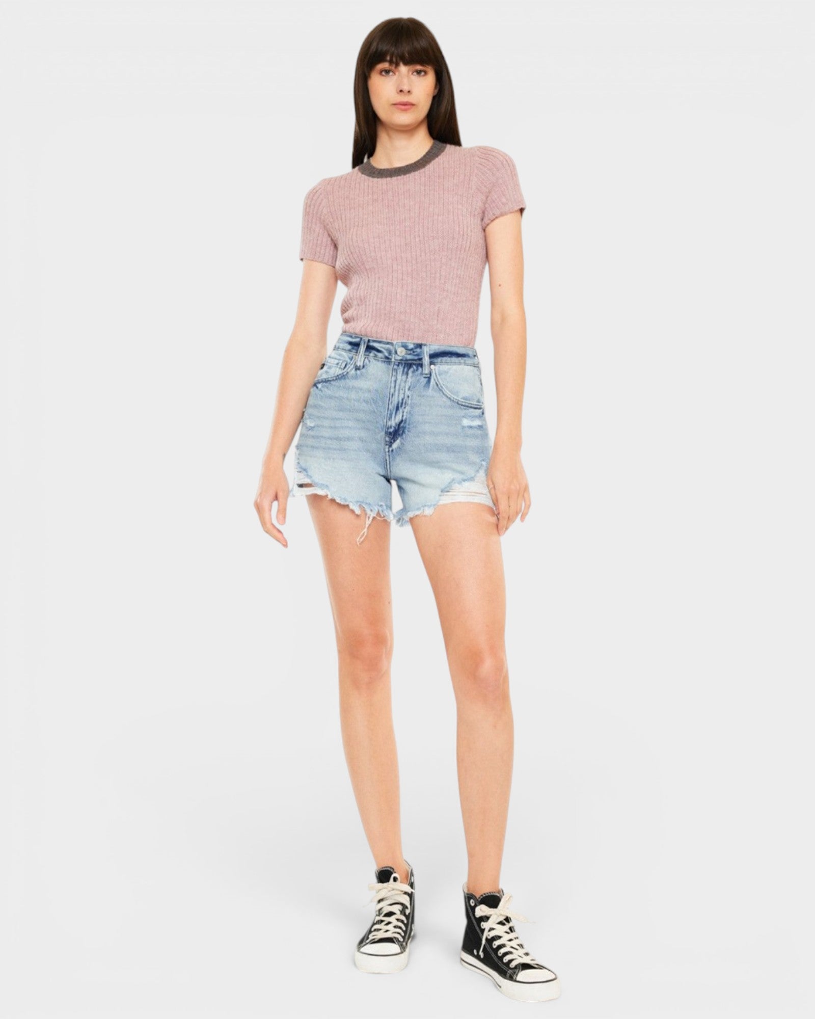 A model wearing a pink short-sleeve knit top, light-wash distressed denim shorts, and black canvas sneakers, standing against a light gray studio background.