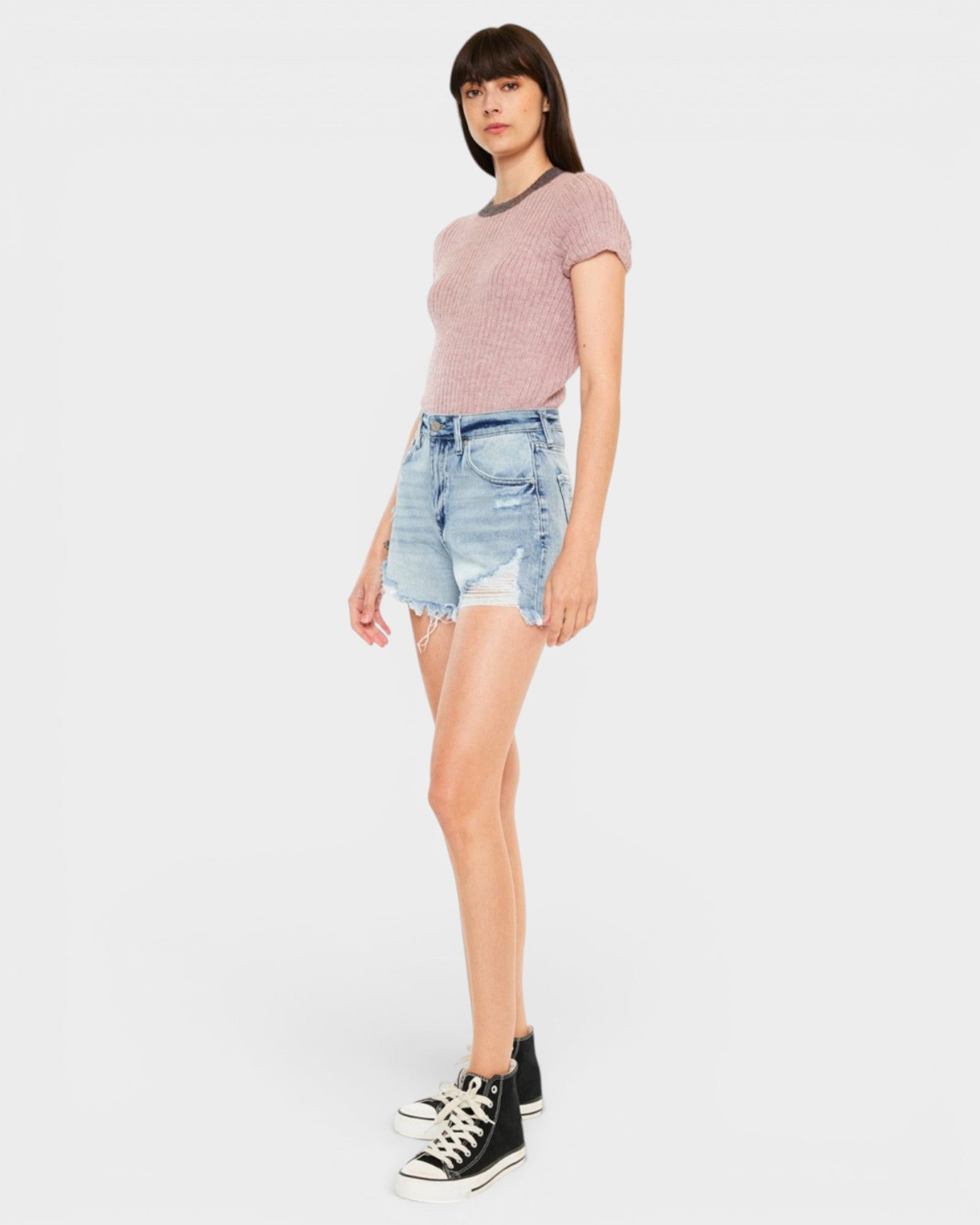 A model wearing a dusty pink short-sleeve knit top, light-wash denim shorts, and black high-top sneakers, standing against a solid light gray background.
