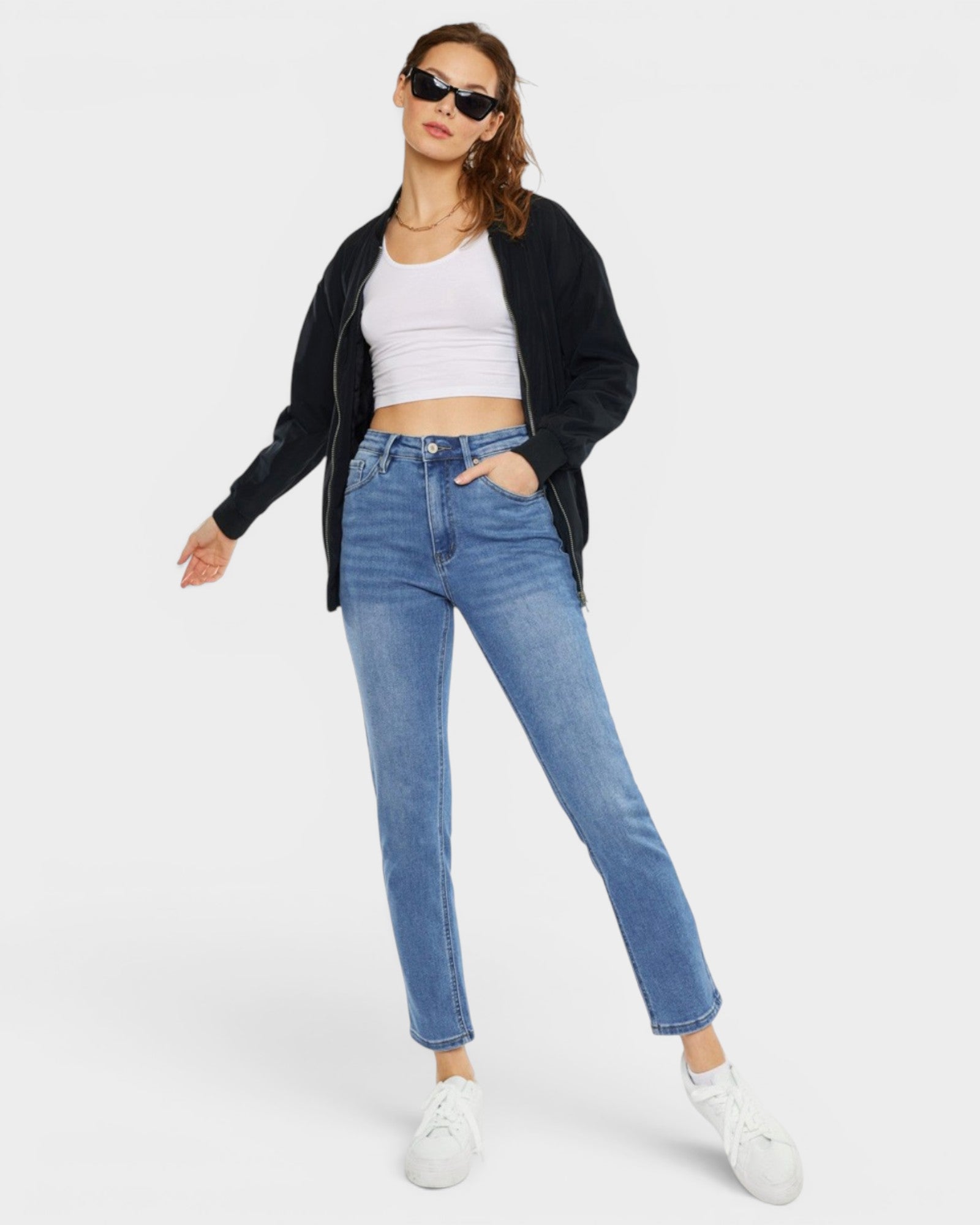 Woman wearing a black jacket, white crop top, blue jeans, and sunglasses on a white background