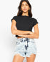 A woman modeling a black short-sleeve crop top with ruffled edges and light-wash distressed denim shorts against a plain white studio background.