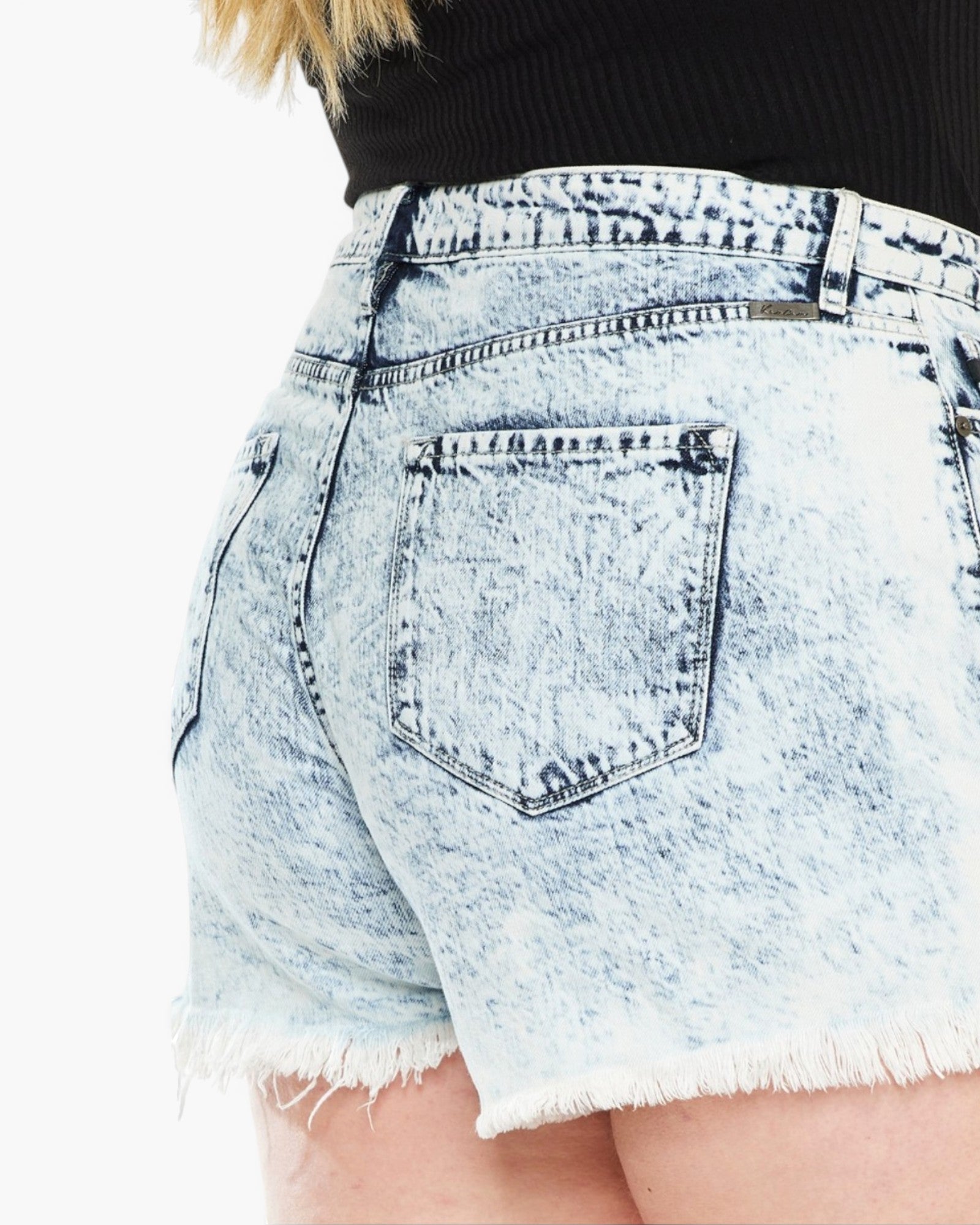 Close-up of the back of light-wash acid-wash denim shorts with a frayed hem, worn with a black ribbed top against a white background.