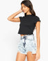 A woman modeling a black short-sleeve ribbed crop top and light-wash distressed denim shorts against a plain white studio background.