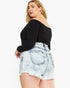 A plus-size model wearing a black off-the-shoulder long-sleeve top and light-wash frayed denim shorts, photographed against a plain white studio background.