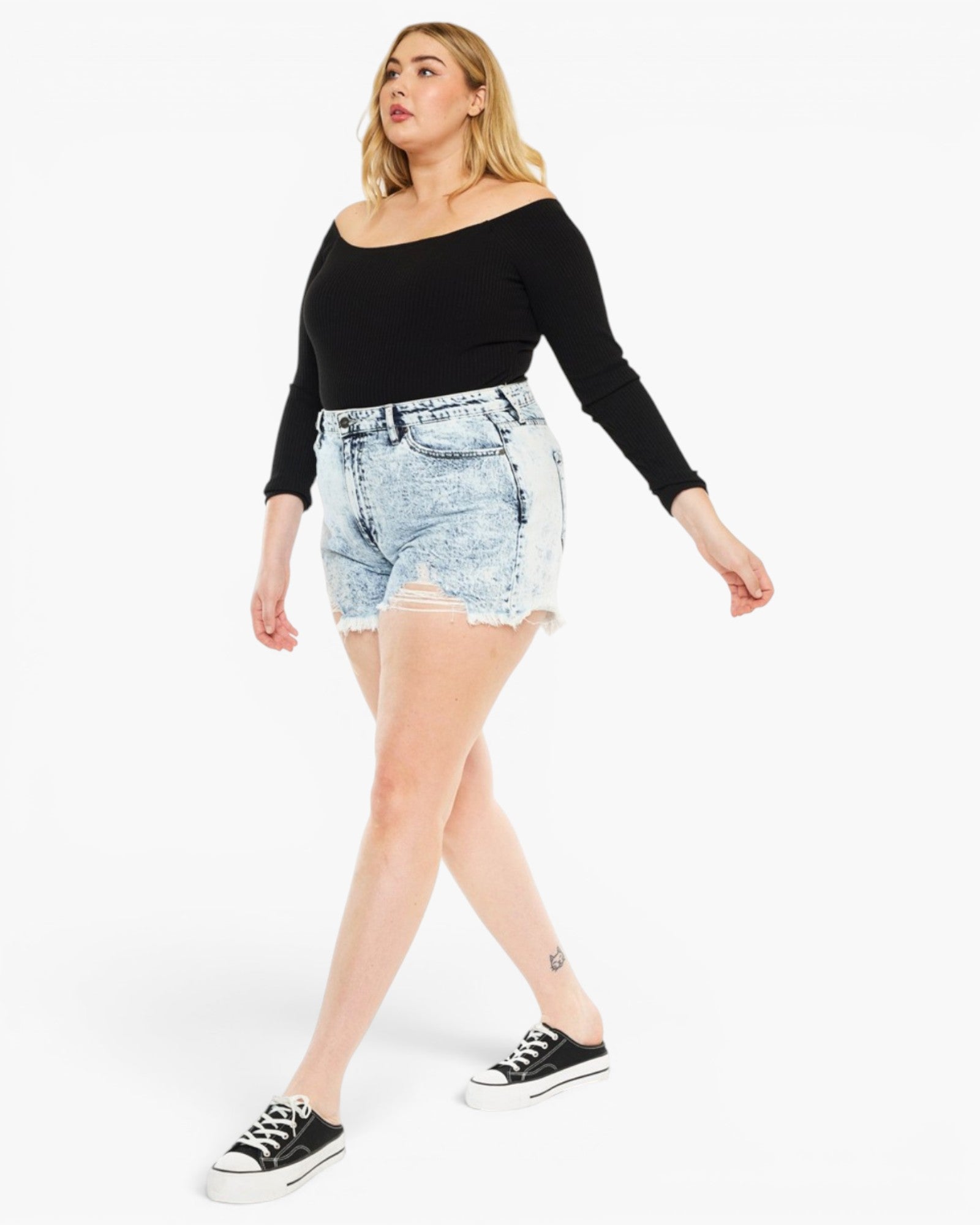 A woman modeling a black off-the-shoulder long-sleeve top and light-wash denim shorts with black low-top sneakers, photographed against a plain white studio background.