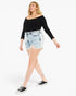 A woman modeling a black off-the-shoulder long-sleeve top and light-wash denim shorts with black low-top sneakers, photographed against a plain white studio background.