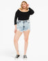 A woman modeling a black off-the-shoulder long-sleeve top and light-wash distressed denim shorts with black low-top sneakers, photographed against a plain white studio background.