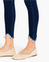 Close-up of a person's legs wearing dark blue frayed-hem skinny jeans and beige flat shoes against a light gray background.