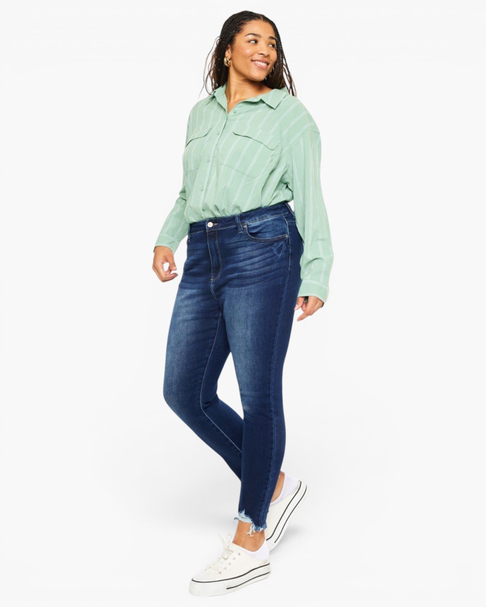 A woman modeling dark blue skinny jeans with a light green striped button-up shirt and white sneakers, standing against a plain white studio background.