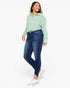 A woman modeling dark blue skinny jeans with a light green striped button-up shirt and white sneakers, standing against a plain white studio background.