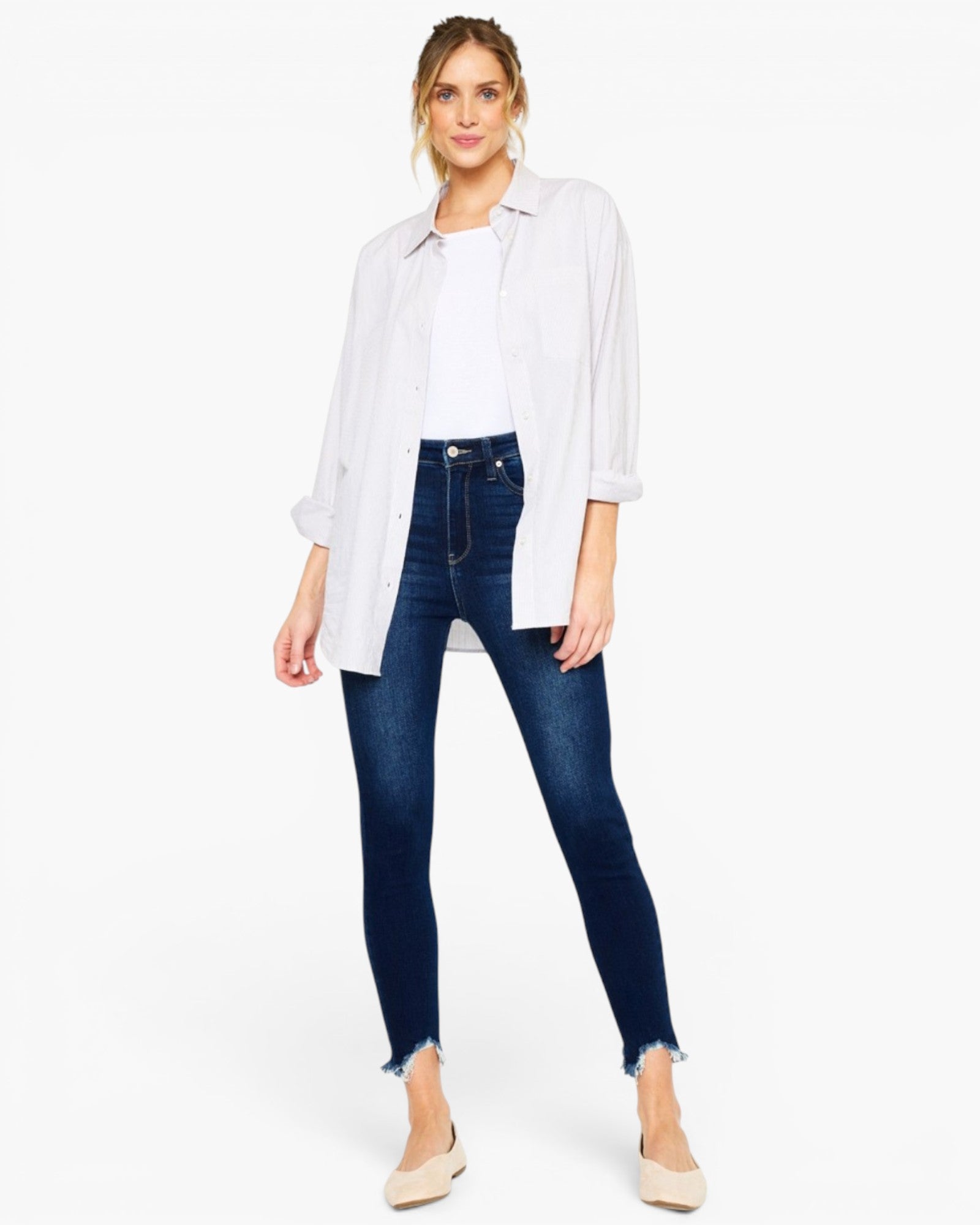 A woman modeling a white button-down shirt layered over a white top, paired with dark blue skinny jeans and beige pointed-toe flats, against a plain white studio background.