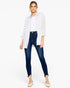 A woman modeling a white button-down shirt layered over a white top, paired with dark blue skinny jeans and beige pointed-toe flats, against a plain white studio background.