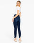 A woman modeling dark wash skinny jeans from a rear three-quarter angle, paired with a white t-shirt and beige flat shoes, against a solid white background.