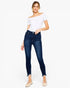 A woman modeling dark blue high-waisted skinny jeans with a frayed hem, paired with a white off-the-shoulder top and beige flats, against a plain white background.