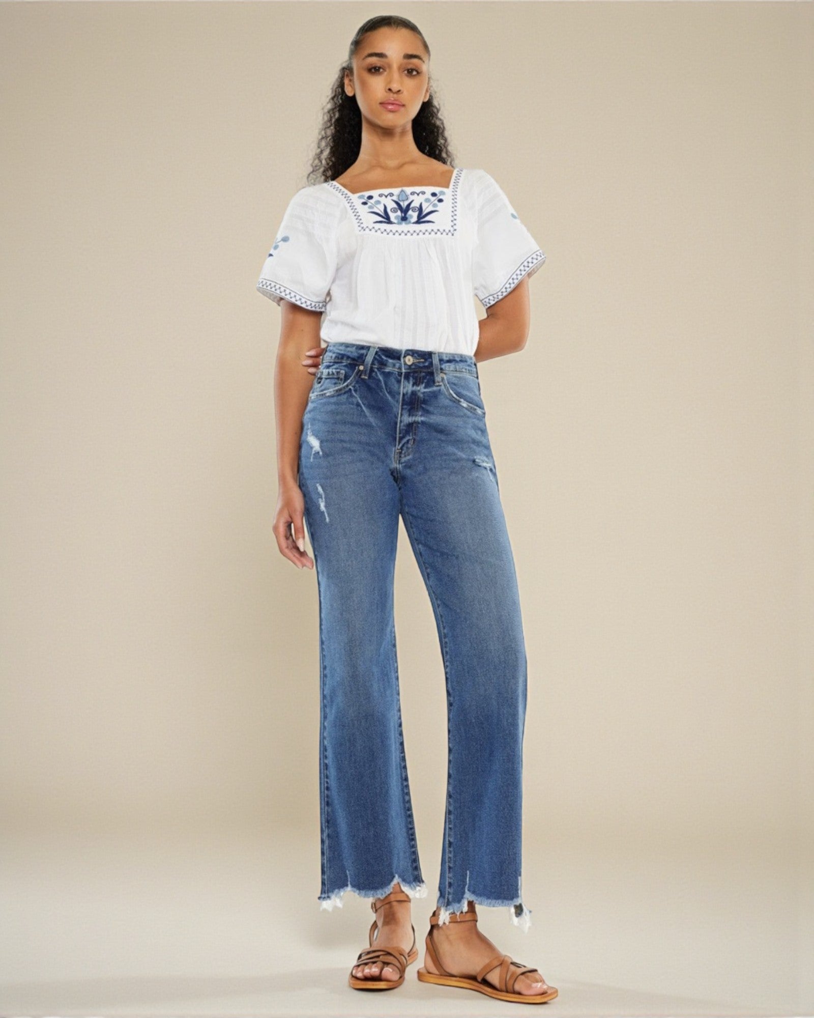 Woman wearing a white embroidered top and blue jeans on a beige background