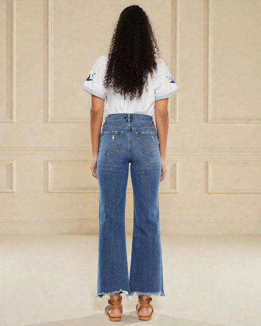 Person wearing blue jeans and a white top with black patterns, standing against a beige paneled wall.