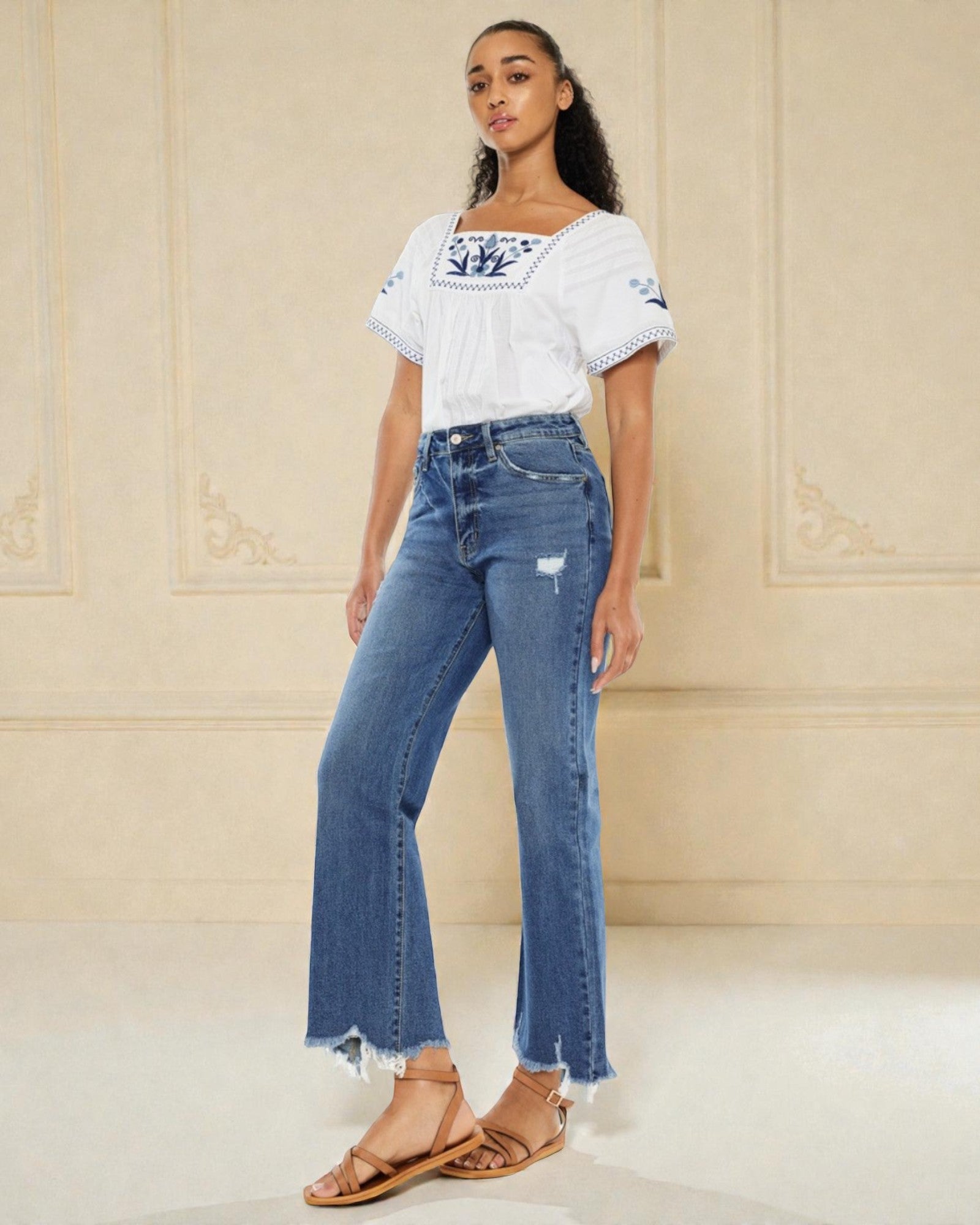 Woman wearing a white blouse with blue patterns and blue jeans against a beige wall.