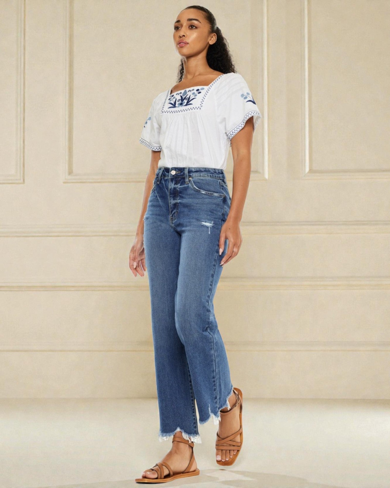 Woman wearing a white embroidered top and blue jeans against a beige wall.