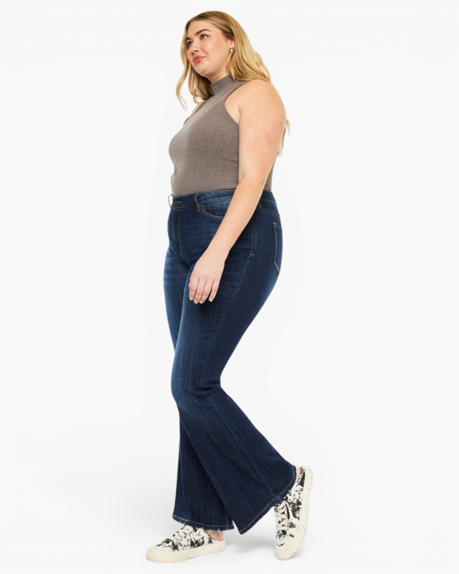 A woman modeling dark blue flare jeans with a ribbed mock neck top and white sneakers, shown from a side profile against a plain white background.