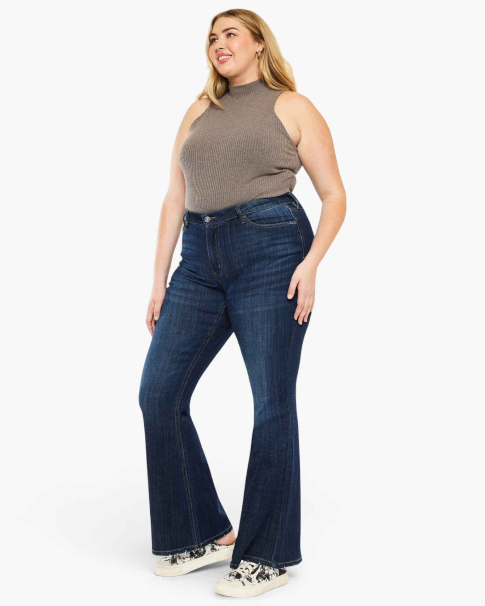A woman modeling dark blue high-waisted flare jeans with a ribbed taupe sleeveless top and white sneakers, standing against a solid white studio background.