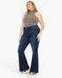 A woman modeling dark blue high-waisted flare jeans with a ribbed taupe sleeveless top and white sneakers, standing against a solid white studio background.