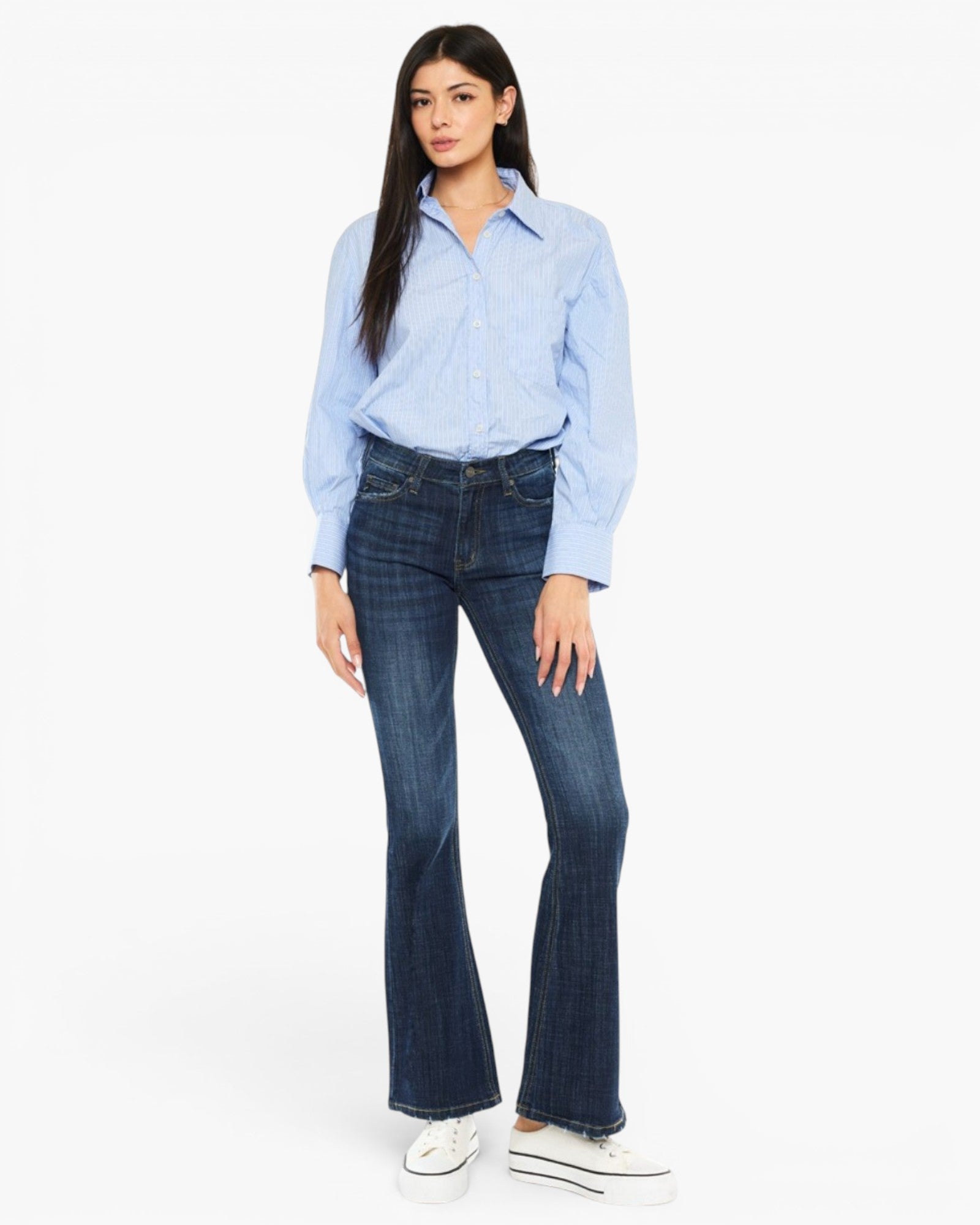 A woman modeling a light blue long-sleeve button-up shirt and dark blue flare jeans with white sneakers, photographed against a plain white studio background.