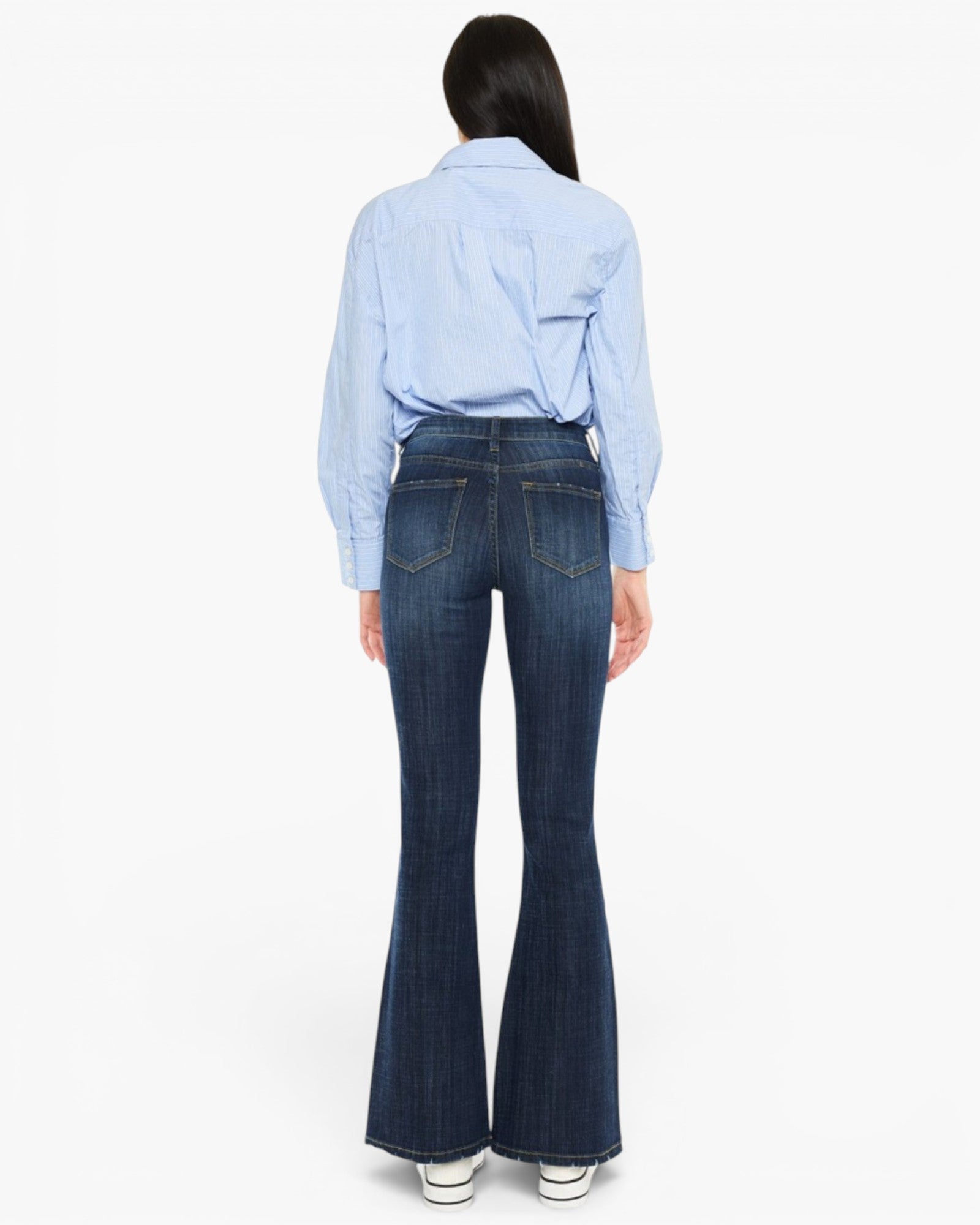 A woman shown from the back wearing a light blue button-up shirt and dark blue flare jeans, standing against a plain white studio background.