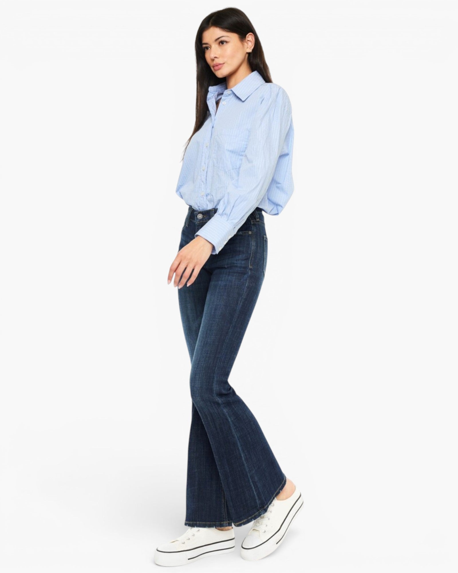 A woman modeling a light blue button-up shirt and dark blue flare jeans with white platform sneakers, photographed against a plain white studio background.