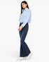 A woman modeling a light blue button-up shirt and dark blue flare jeans with white platform sneakers, photographed against a plain white studio background.
