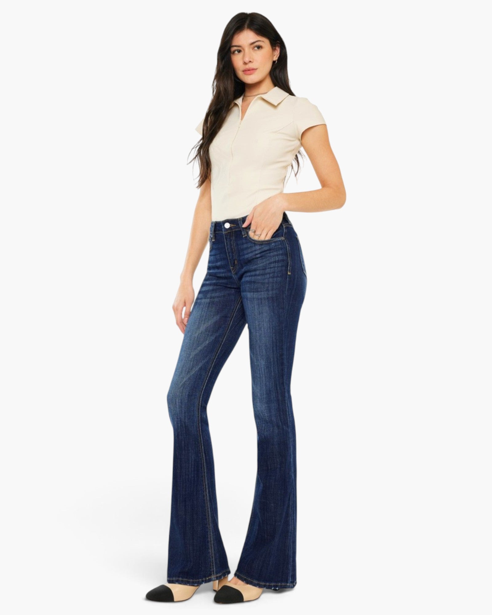 A woman modeling a pair of dark blue high-waisted flare jeans, paired with a cream short-sleeve collared shirt and black-and-white flats, against a plain white studio background.
