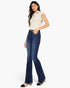 A woman modeling a pair of dark blue high-waisted flare jeans with a cream short-sleeve collared top, standing against a plain white studio background.