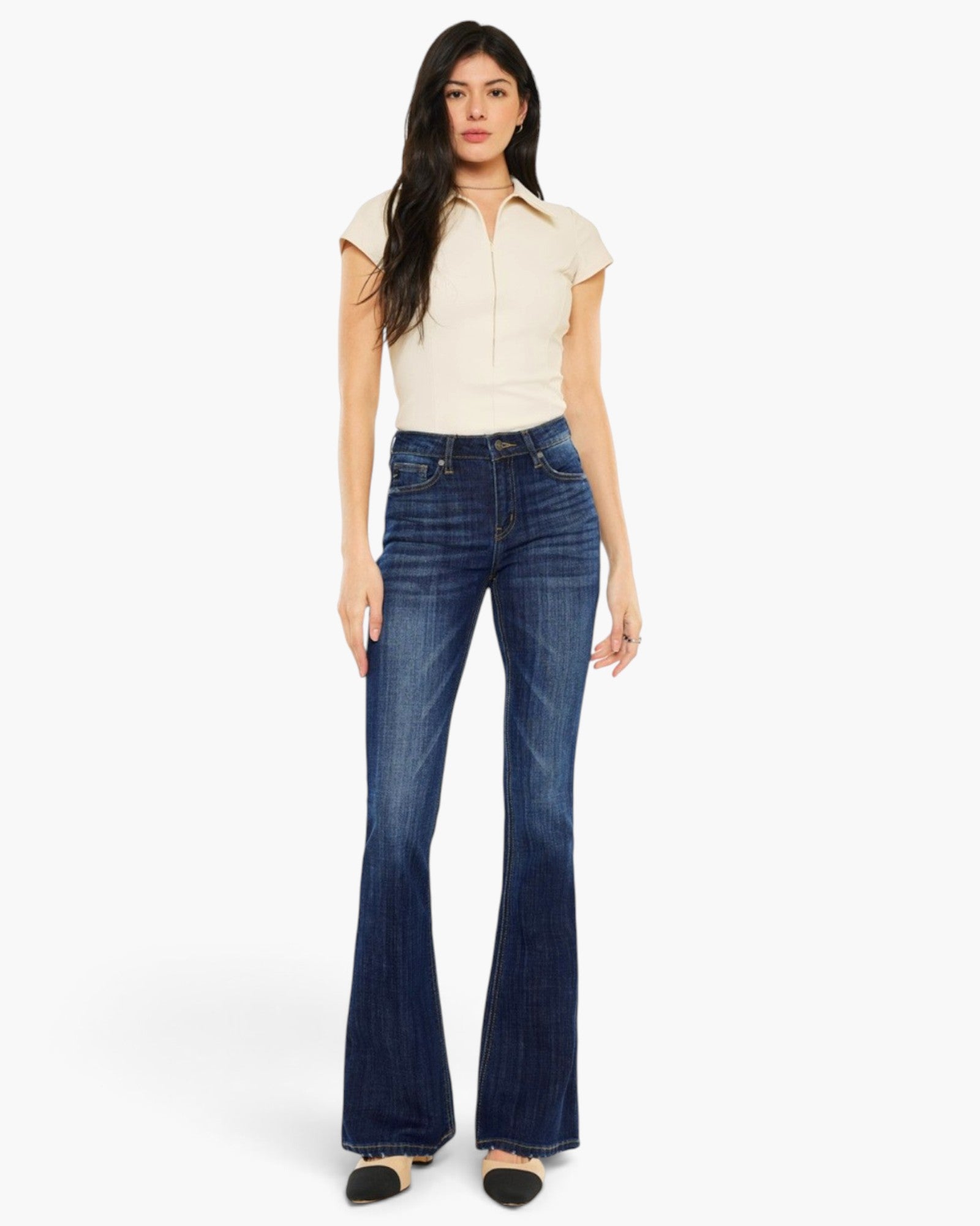 A woman modeling a pair of dark blue high-waisted flare jeans with a cream short-sleeve collared top, standing against a plain white studio background.