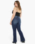 A woman modeling a pair of dark blue flare jeans from the back, paired with a black spaghetti-strap top and black heels, against a plain white studio background.