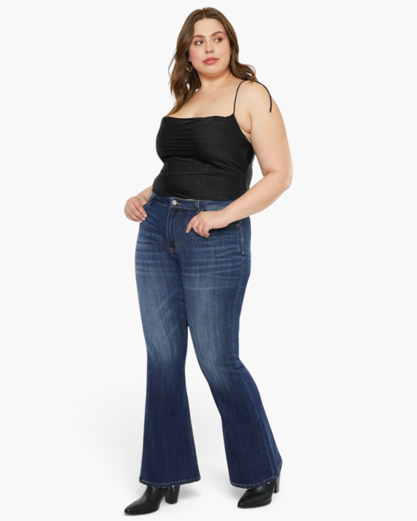 A woman modeling a pair of dark blue flare jeans with a black spaghetti-strap top, standing against a plain white studio background.