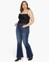 A woman modeling a pair of dark blue flare jeans with a black spaghetti-strap top, standing against a plain white studio background.
