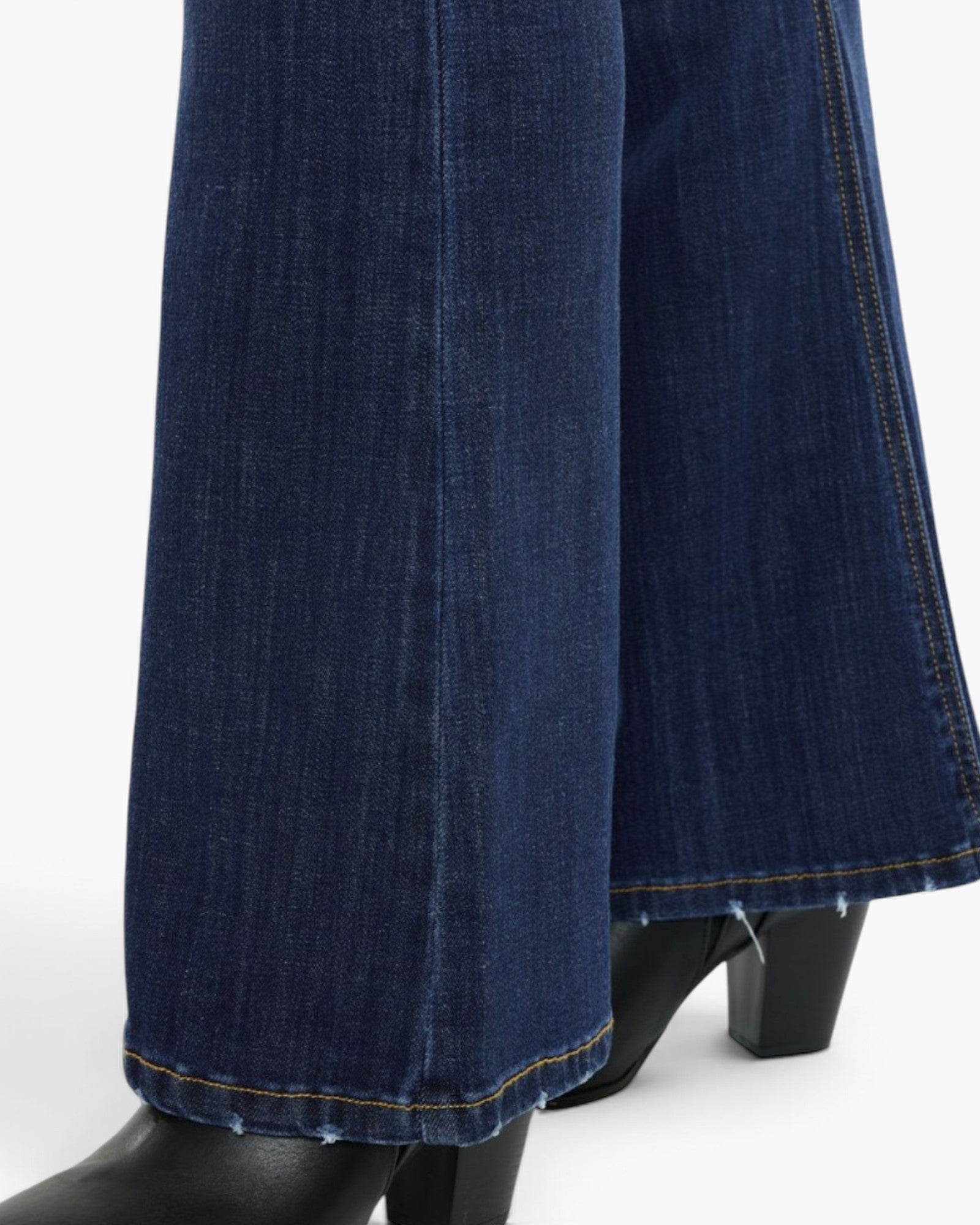 Close-up of dark blue denim jeans with a wide leg and raw frayed hems, shown on a mannequin with black block-heel boots against a white studio background.