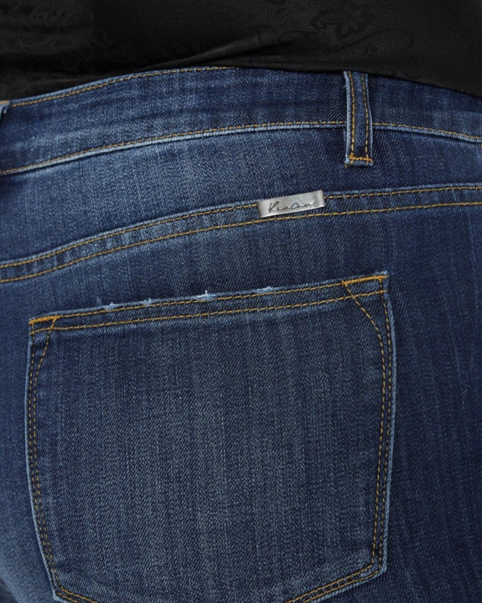 Close-up of the back pocket of dark blue denim jeans with yellow contrast stitching and a small metal brand tag reading 'Kancan'.