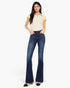 A woman modeling a pair of dark blue flared jeans with a cream short-sleeve collared top, standing against a plain white studio background.