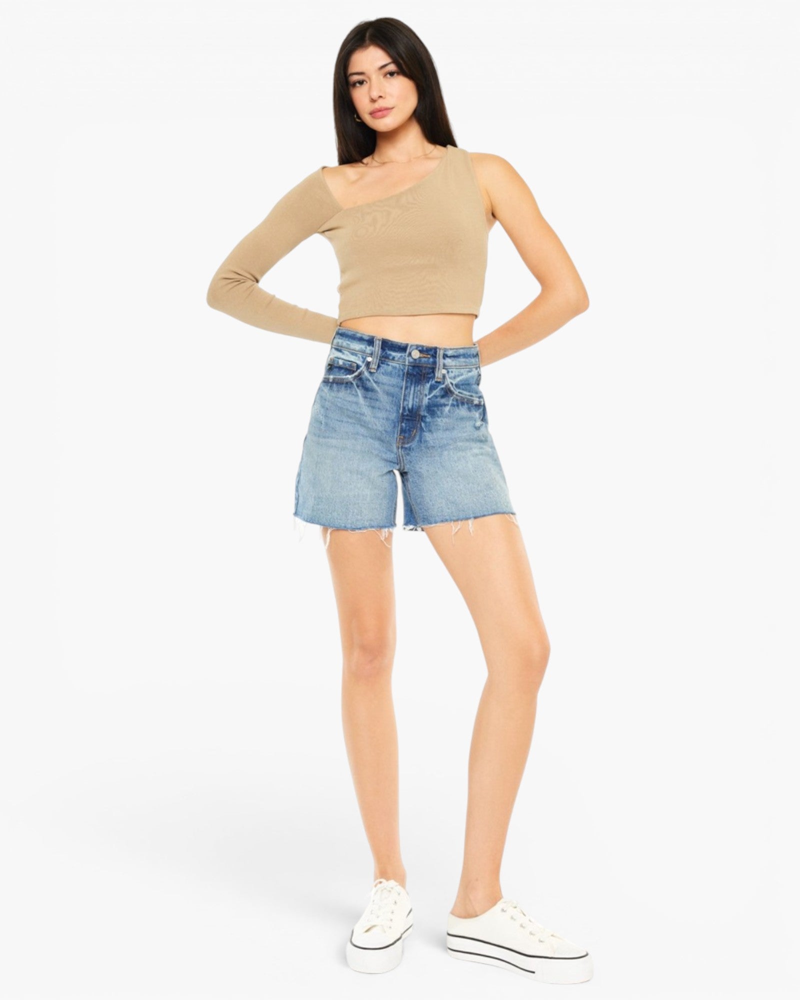 A woman modeling a beige long-sleeve crop top and light-wash blue denim shorts against a plain light gray studio background.