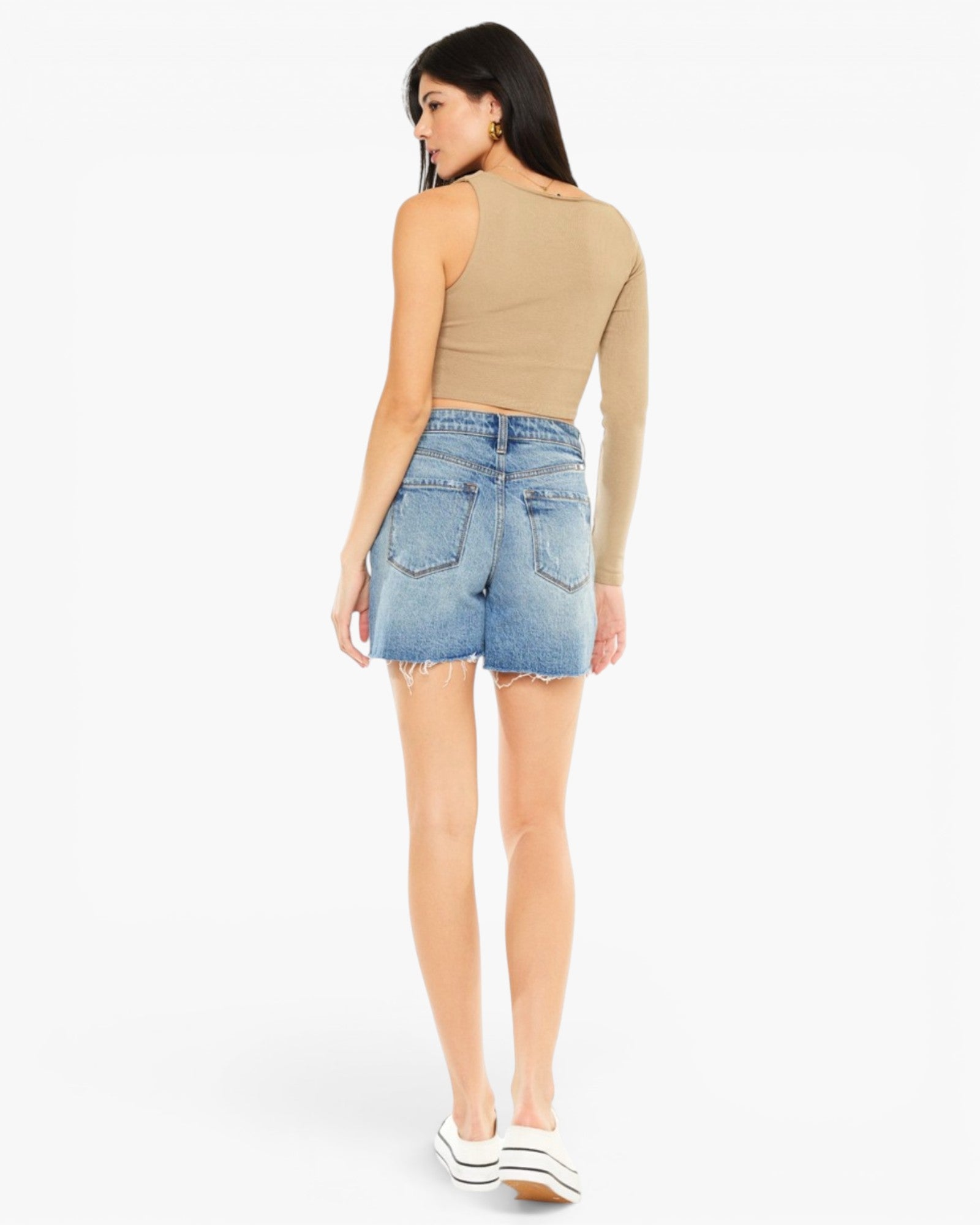 A woman shown from the back wearing a beige long-sleeve crop top and light-wash denim shorts, paired with white platform sneakers, against a plain white studio background.