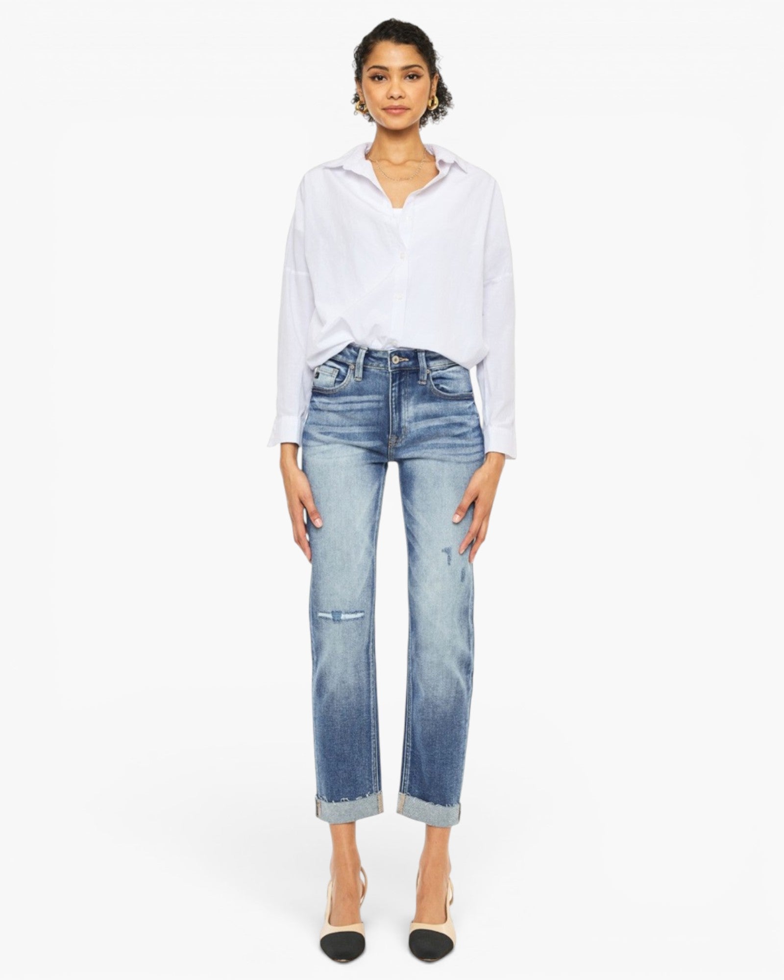 A model stands against a plain white studio background wearing a white long-sleeve button-up shirt, light-wash blue cropped jeans with faded details and rolled cuffs, and black pointed-toe heels.