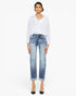 A model stands against a plain white studio background wearing a white long-sleeve button-up shirt, light-wash blue cropped jeans with faded details and rolled cuffs, and black pointed-toe heels.
