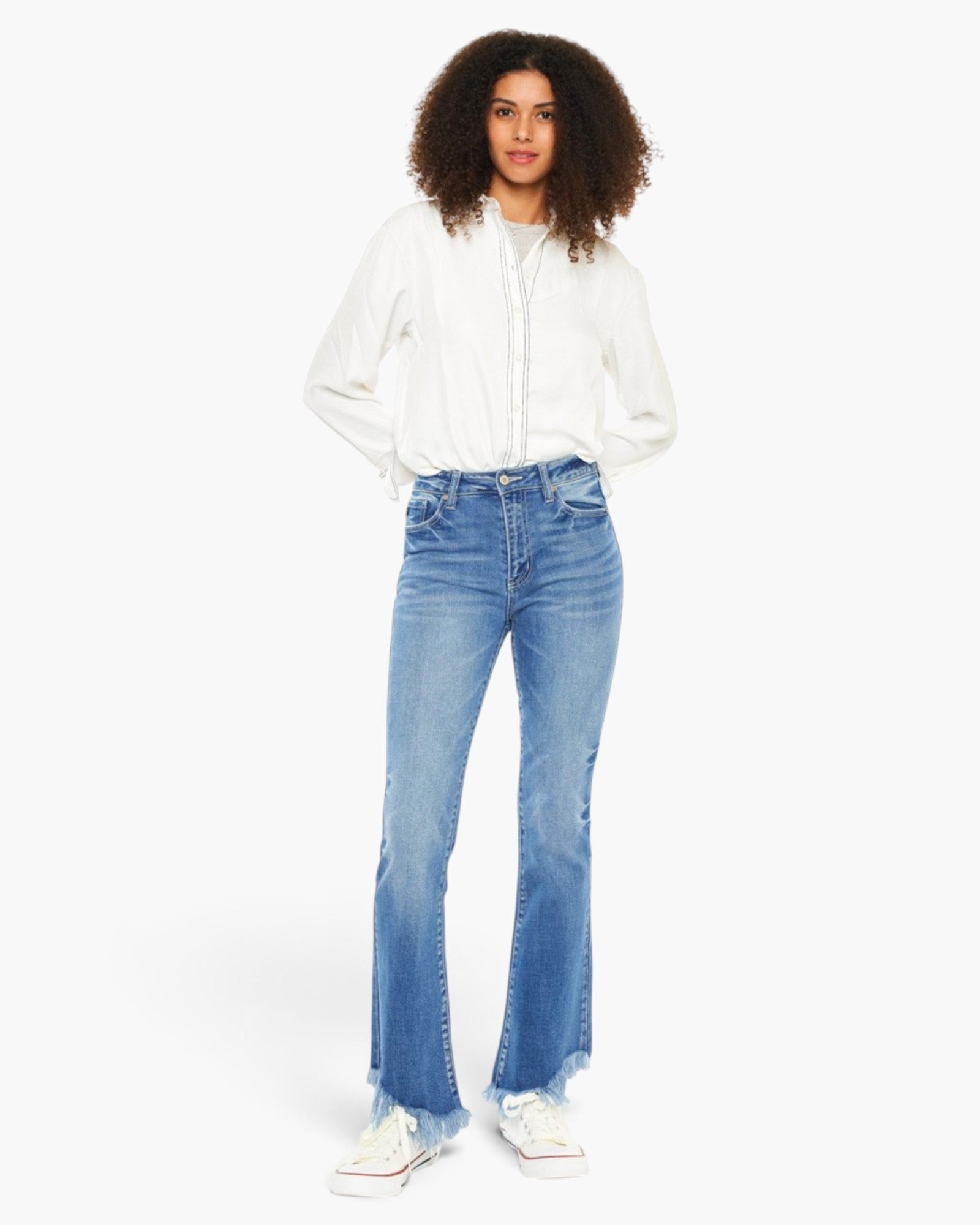 A model wearing a white long-sleeve button-up shirt and light blue high-waisted flare jeans with raw hems, paired with white sneakers, against a plain white studio background.