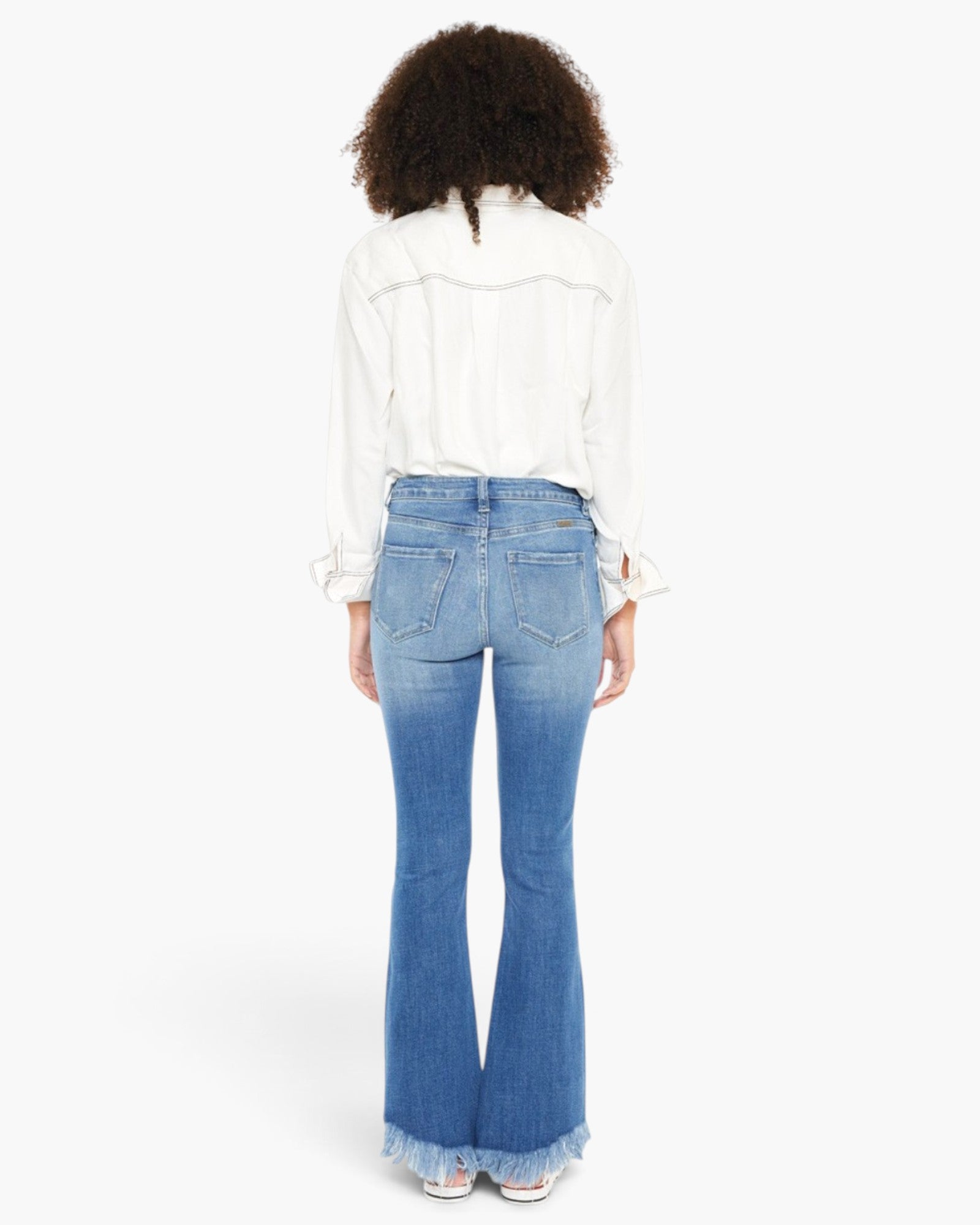 A woman viewed from the back wearing a white long-sleeve blouse and light blue flared jeans with a frayed hem, against a plain white studio background.