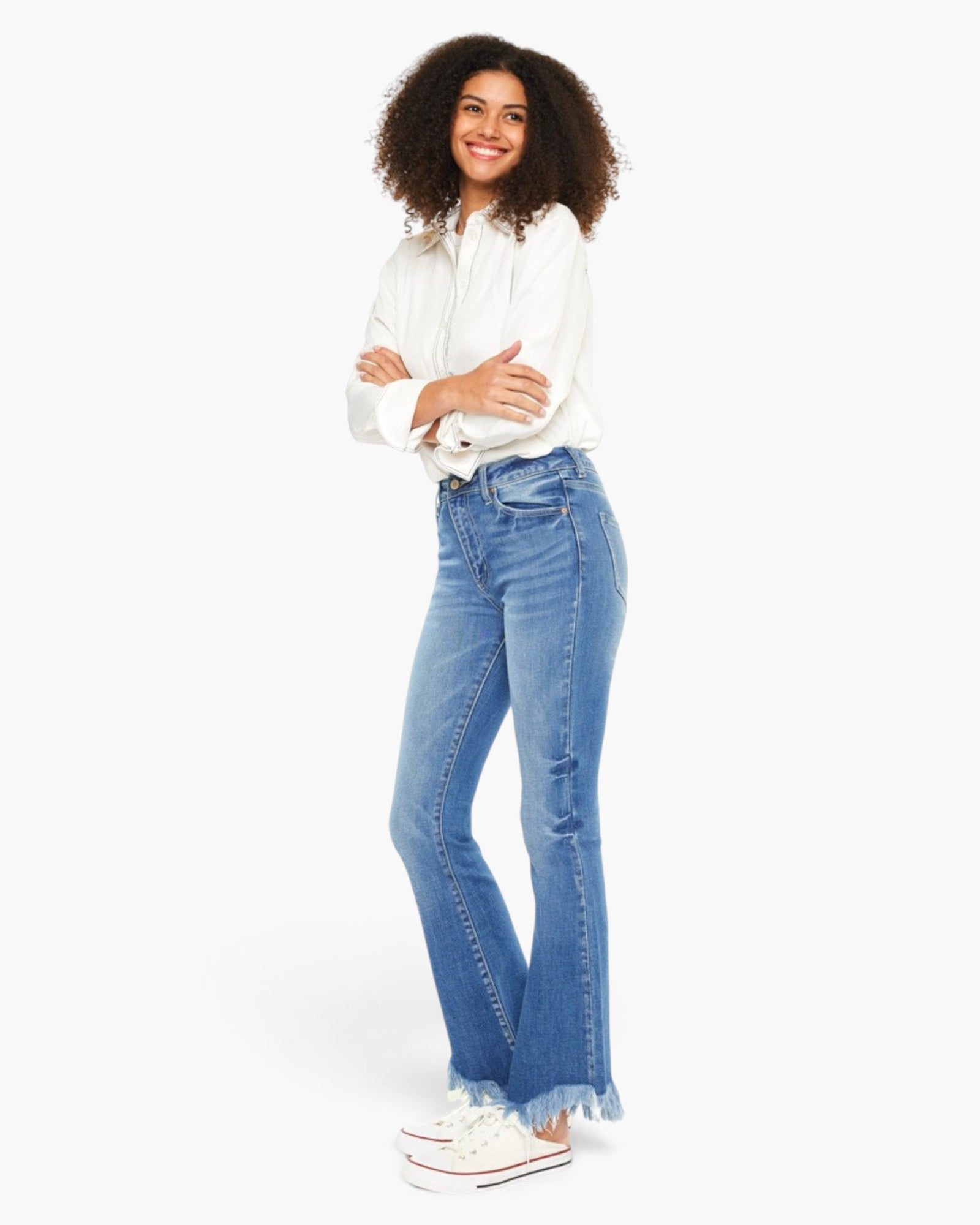 A woman modeling a pair of light blue high-waisted flare jeans with a frayed hem, paired with a white button-down shirt and white sneakers, against a plain white studio background.