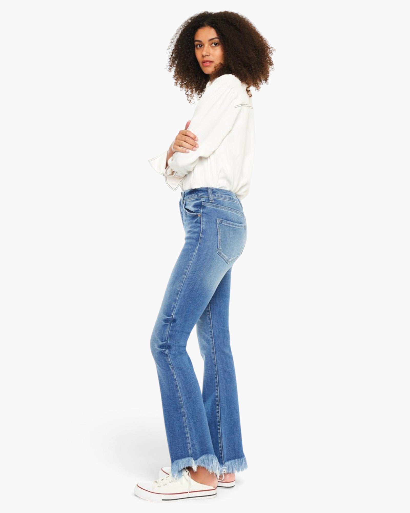 A woman modeling a pair of light blue flared jeans with a frayed hem, paired with a white long-sleeve shirt and white sneakers, shown from a side-back angle against a plain white background.