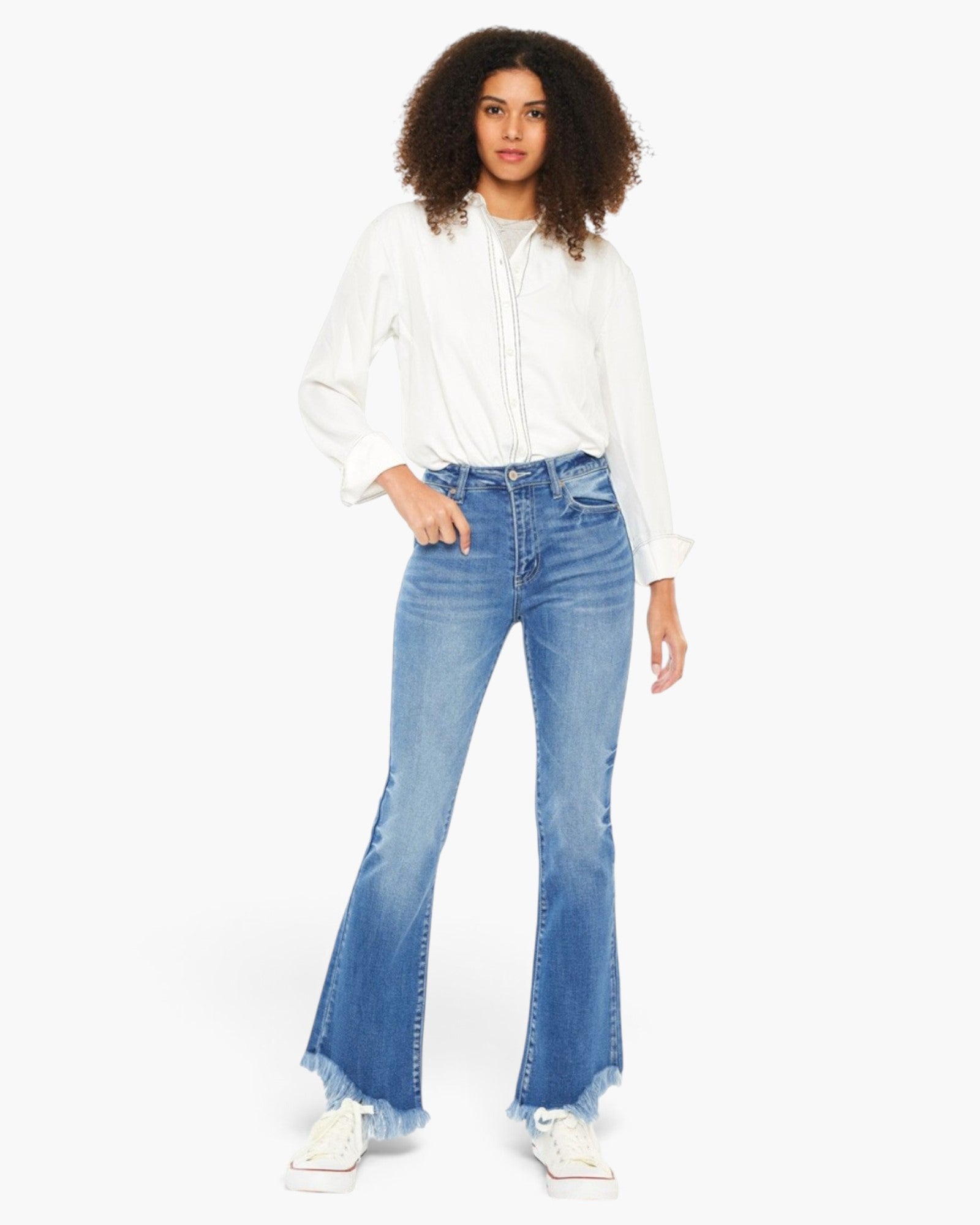 A model wearing a white long-sleeve button-up shirt and blue high-waisted flare jeans with a raw hem, paired with white sneakers, against a plain white background.