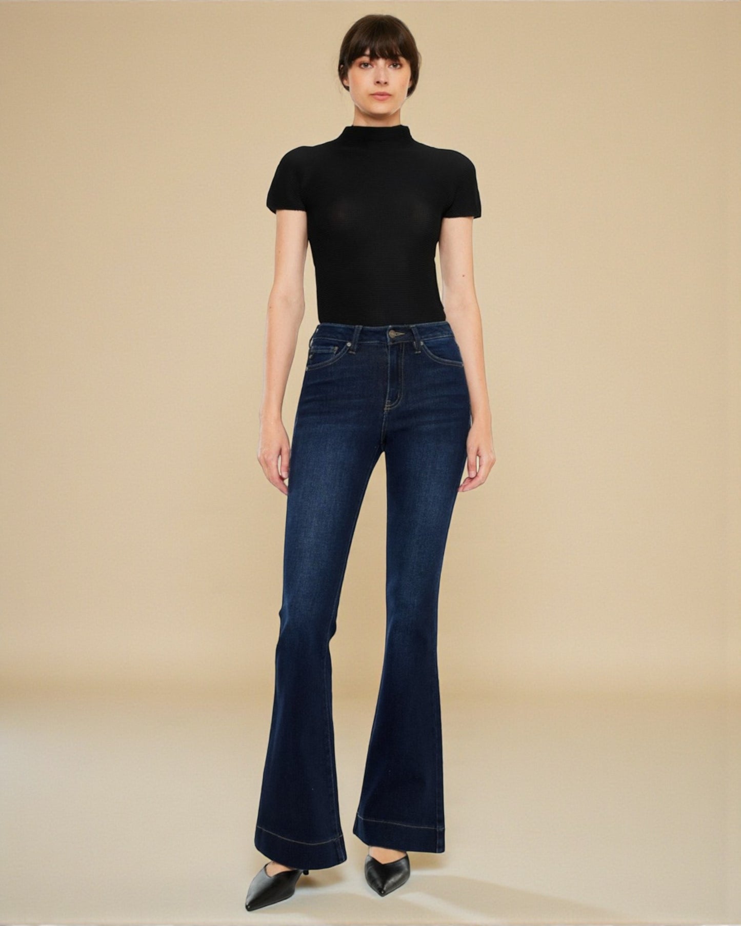 Person wearing dark blue flared jeans and a black turtleneck against a beige background