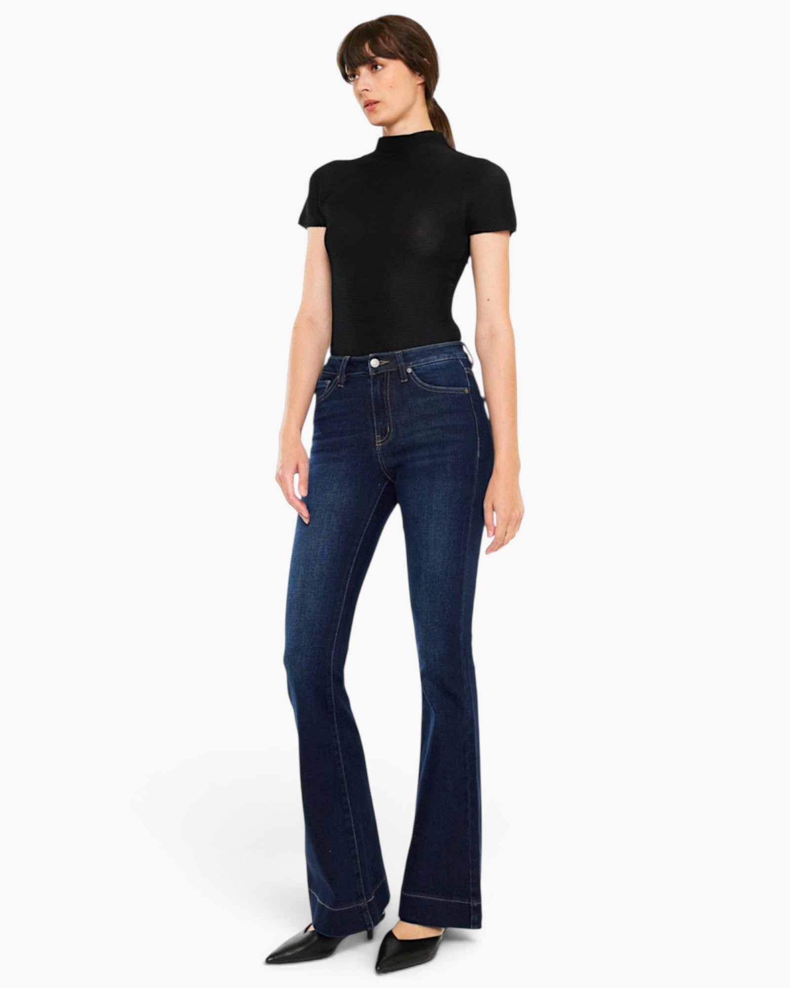 A model wearing a black short-sleeve mock neck top and dark blue high-waisted flare jeans, paired with black pointed flats, against a plain white studio background.