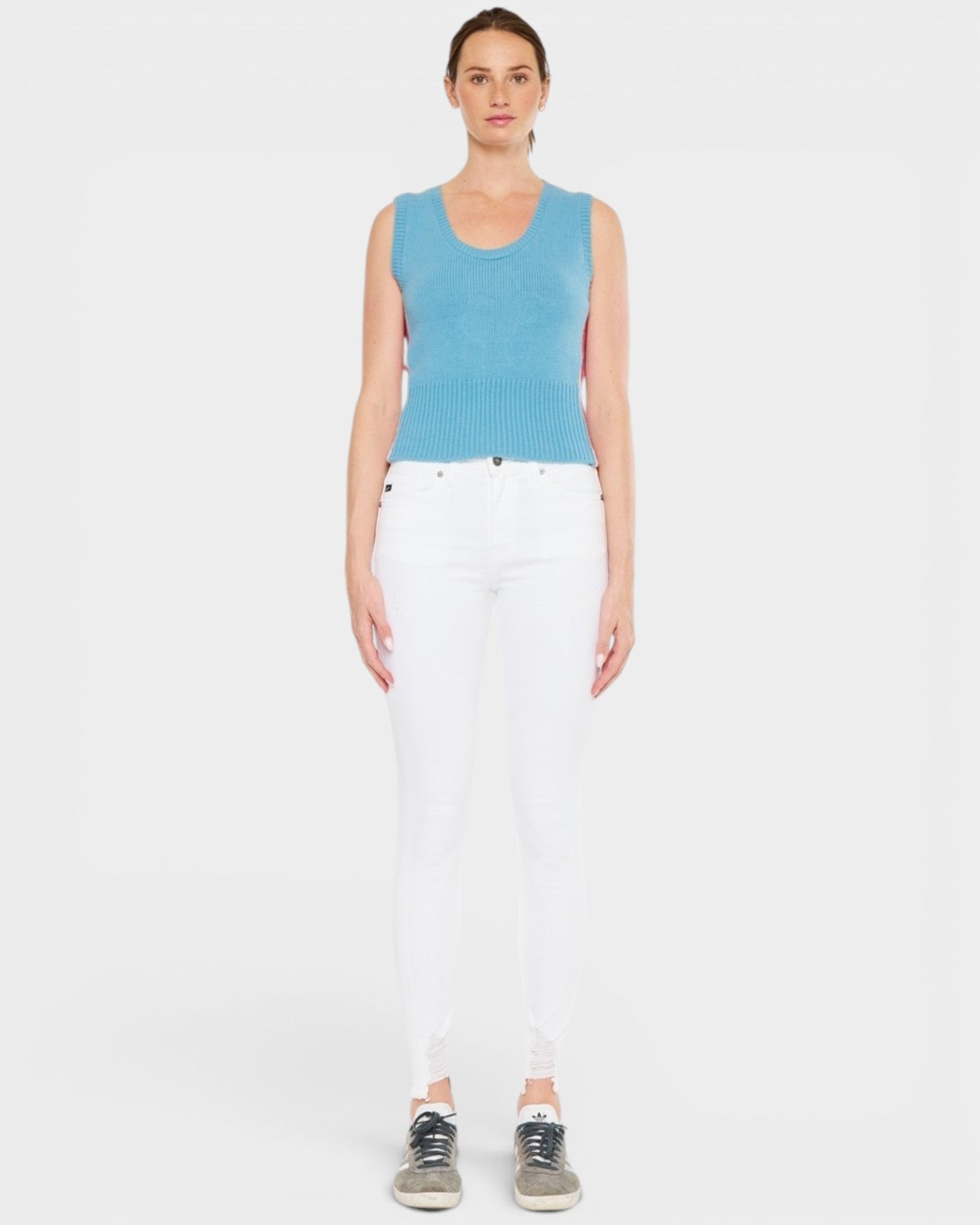Woman wearing a blue sleeveless top and white pants on a plain background